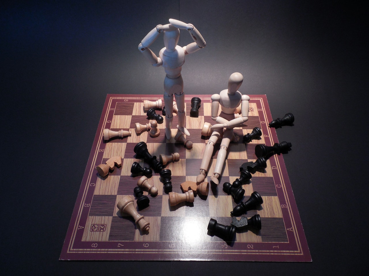 Photo of chess board with all the pieces knocked over and two wooden human figurines with their hands on their head trying to figure out how to overcome their mistakes.